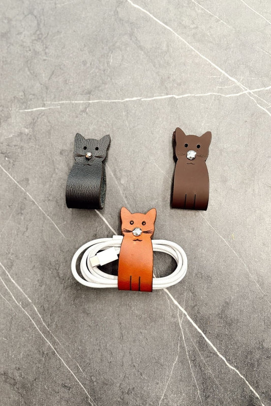 Tres organizadores de cable en cuero con forma de gato, en negro, café y miel; el miel con un cable enrollado