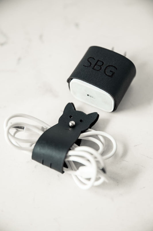 Protector de cargador en cuero negro con unas iniciales grabadas, junto a un organizador de cable negro con forma de gato