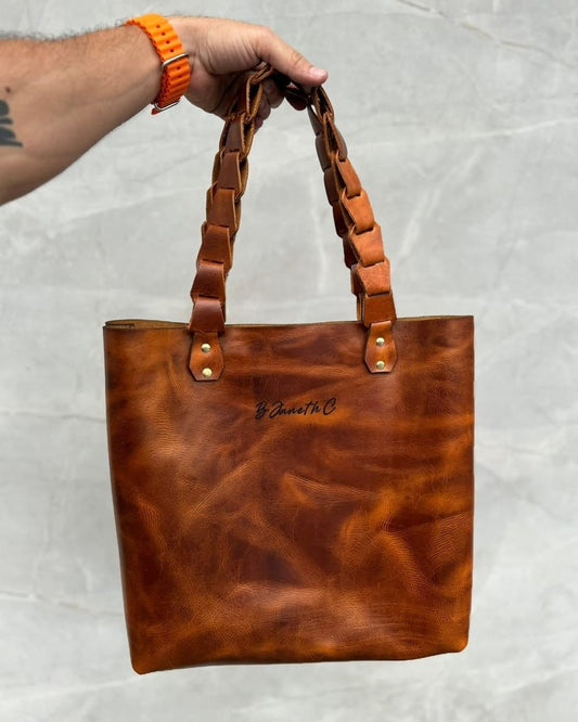 Bolso tote de cuero rústico miel con asas trenzadas y un nombre grabado, en la mano