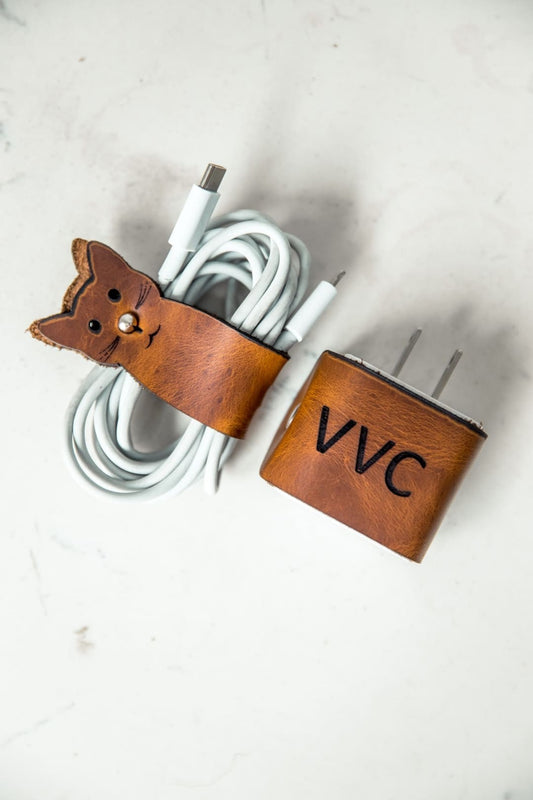Protector de cargador en cuero miel con unas iniciales grabadas, junto a un organizador de cable miel con forma de gato